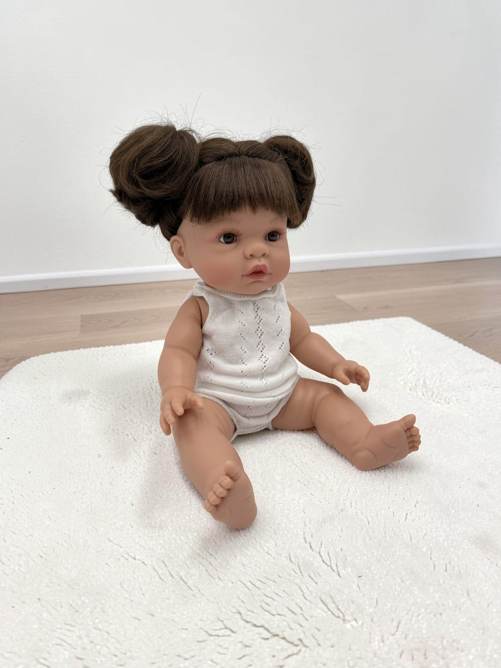 A realistic baby doll with brown hair styled in two buns sits on a white textured rug in a room with light wood flooring and a white wall.