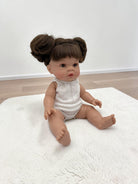 A realistic baby doll with brown hair styled in two buns sits on a white textured rug in a room with light wood flooring and a white wall.