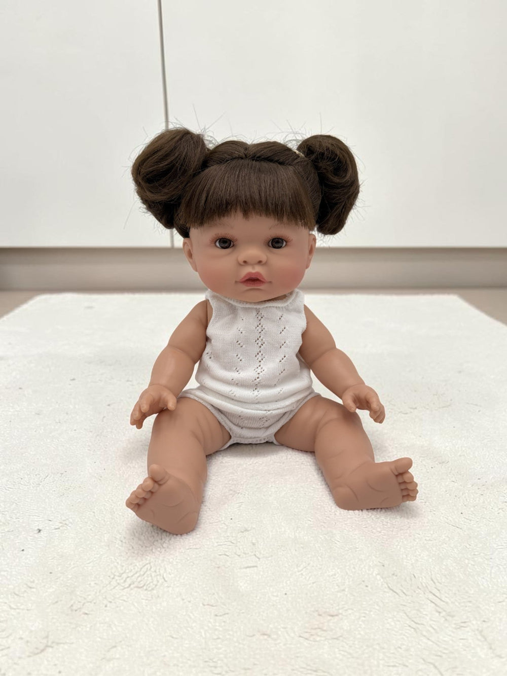 A realistic baby doll with brown hair styled in two buns, wearing a white sleeveless bodysuit, sitting on a white textured rug.