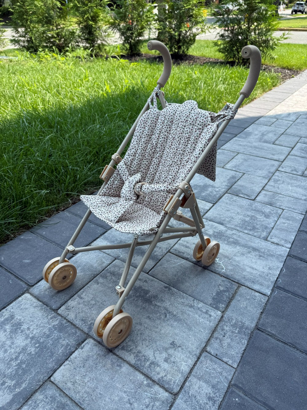 A stroller with a beige frame and a patterned cream and brown fabric seat cover, parked on a gray paved pathway with green grass and trees in the background.