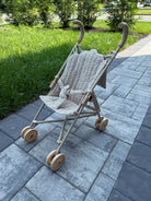 A stroller with a beige frame and a patterned cream and brown fabric seat cover, parked on a gray paved pathway with green grass and trees in the background.