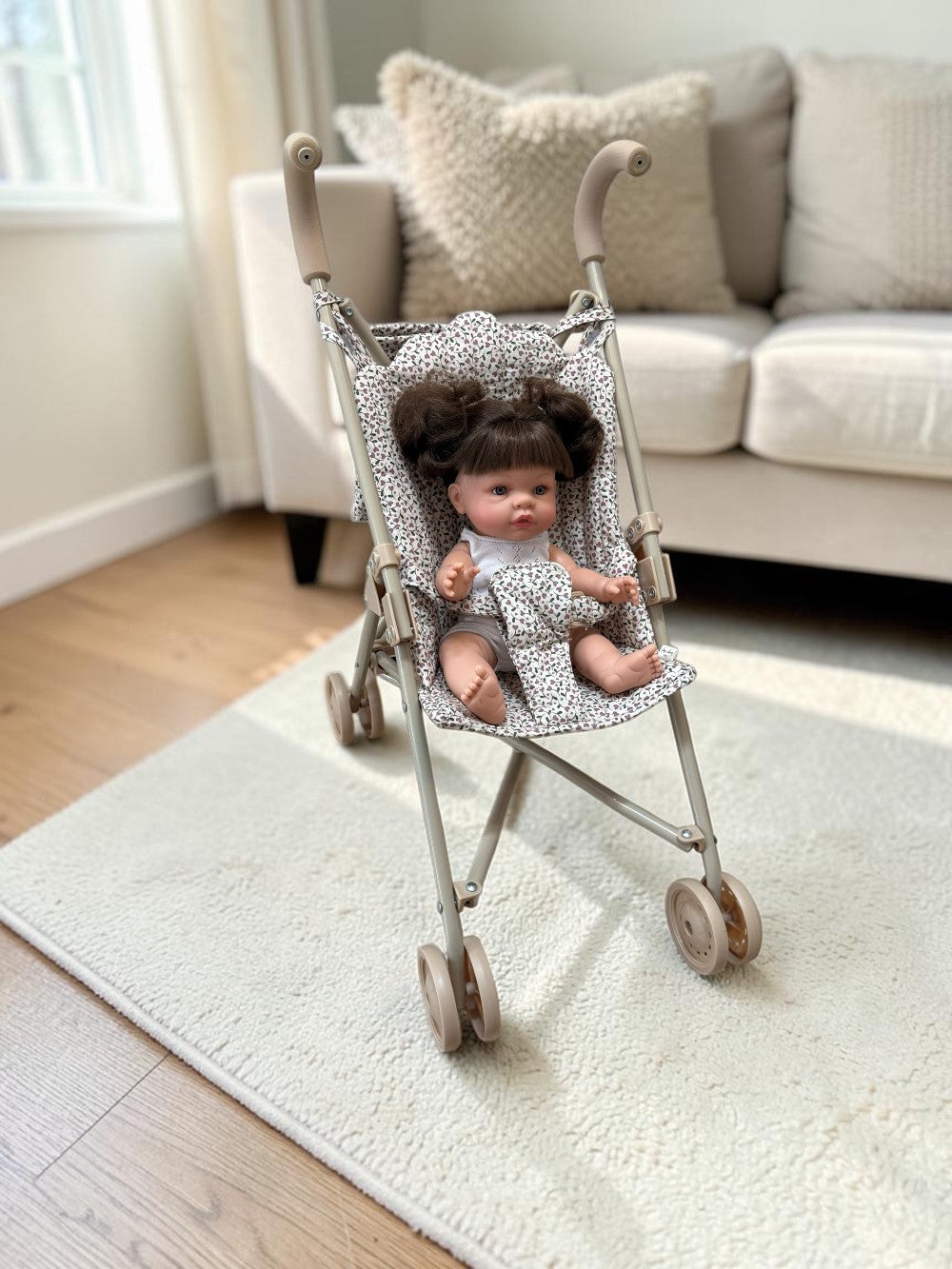 A miniature doll stroller with a gray and white patterned fabric seat, shown on a cream rug in a bright living room with a doll sitting inside.