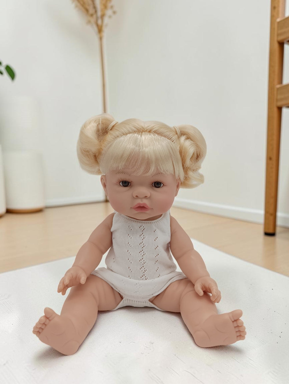 Baby doll with blonde hair styled in pigtails wearing a white onesie on a light surface.