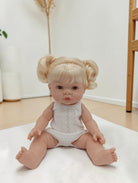 Baby doll with blonde hair styled in pigtails wearing a white onesie on a light surface.