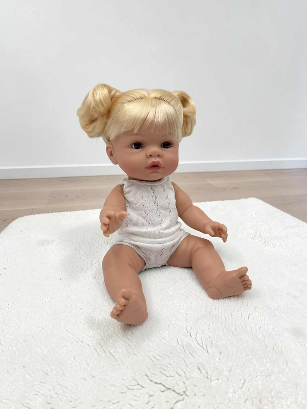 A realistic baby doll with blonde hair in two buns sits on a white textured rug. The doll is wearing a white sleeveless knit romper and is photographed against a plain white wall and light wood floor.