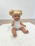 A realistic baby doll with blonde hair in two buns sits on a white textured rug. The doll is wearing a white sleeveless knit romper and is photographed against a plain white wall and light wood floor.