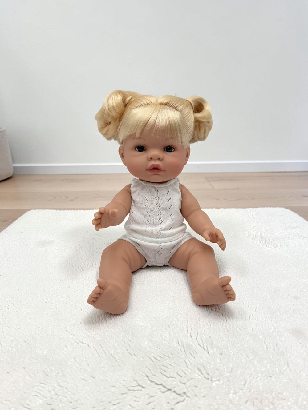 A realistic baby doll with blonde hair in two buns sits on a white textured rug in a room with light wood flooring and a white wall.