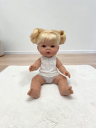 A realistic baby doll with blonde hair in two buns sits on a white textured rug in a room with light wood flooring and a white wall.