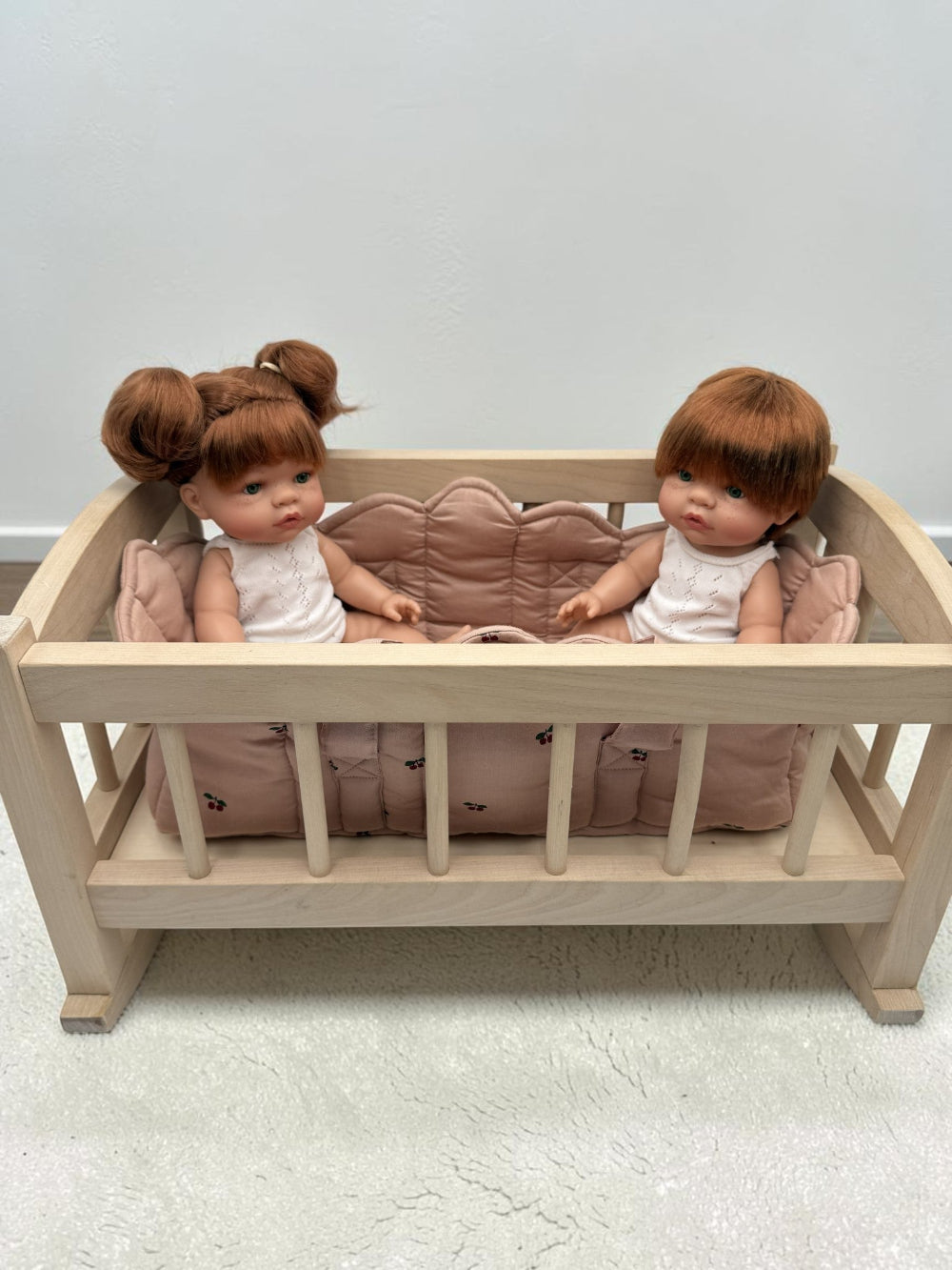 A light natural wood doll bed with a pink quilted cushion, shown with two baby dolls sitting inside against a plain light gray wall and textured floor.
