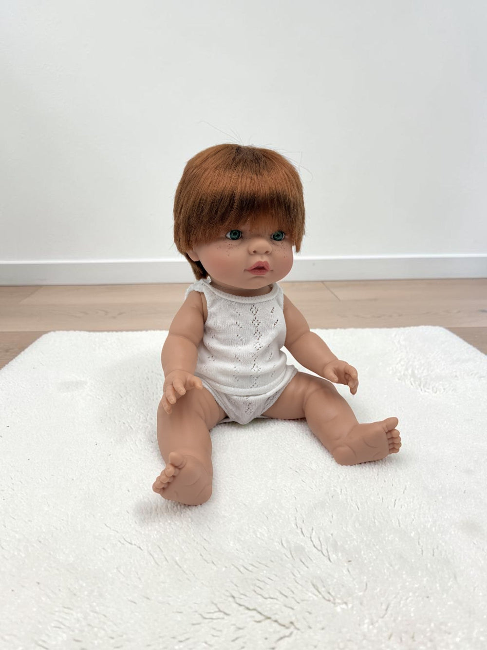 A realistic baby doll with brown hair sits on a white textured rug in a room with light wood flooring and a white wall.