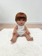A realistic baby doll with short brown hair sits on a white textured rug against a plain white wall and light wood floor.