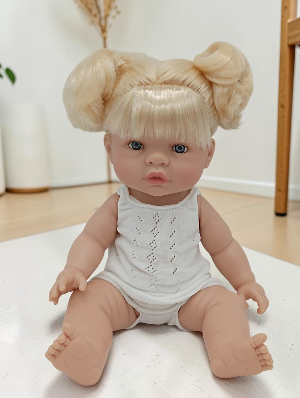 Realistic baby doll with blonde hair styled in buns, wearing a white outfit, on a light-colored surface.