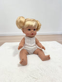 A baby doll with blonde hair styled in two buns sits on a white textured rug against a plain white wall and light wood floor.