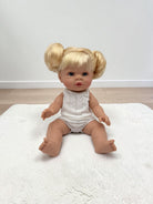 A realistic baby doll with blonde pigtails sits on a white textured rug in front of a plain white wall and light wood floor. The doll is wearing a white sleeveless knit romper.