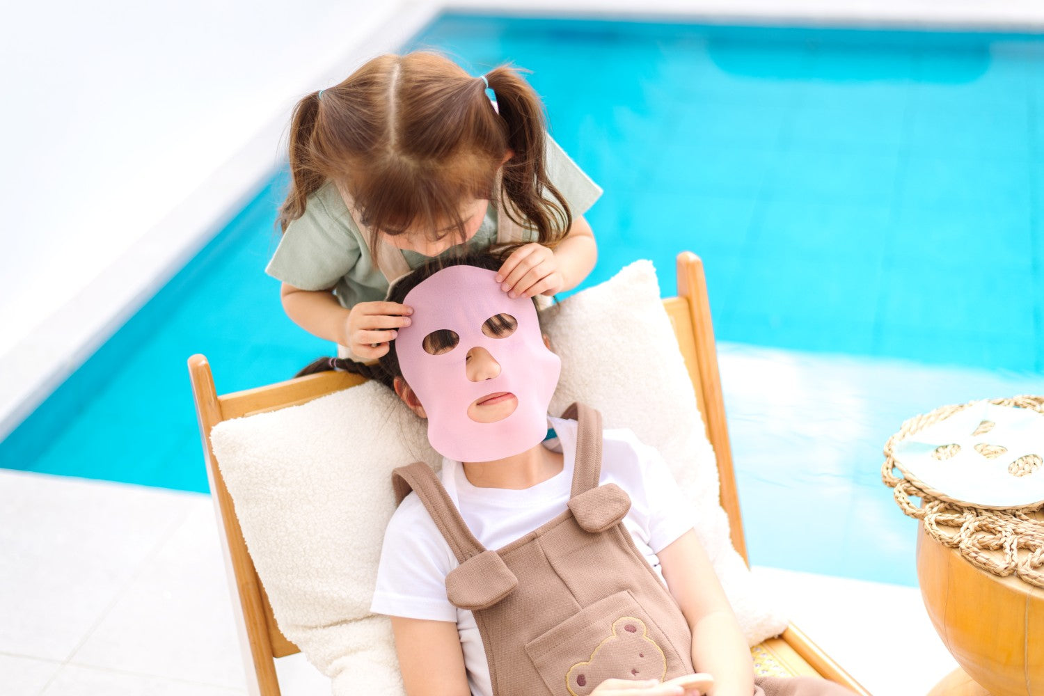 Child applying a face mask to an adult's face by a poolside