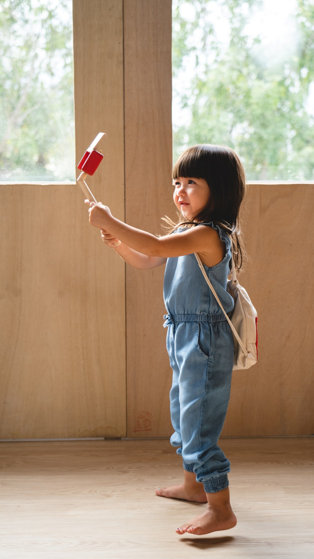 Child in a denim jumpsuit holding a toy in a room with large windows.