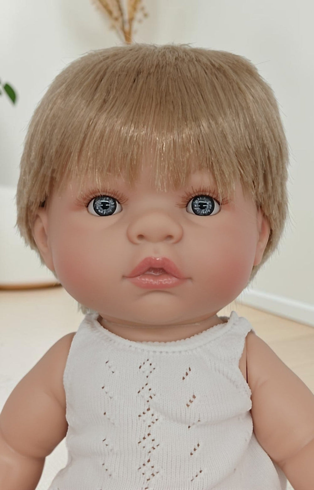 Realistic baby doll with blonde hair and blue eyes wearing a white sleeveless top.