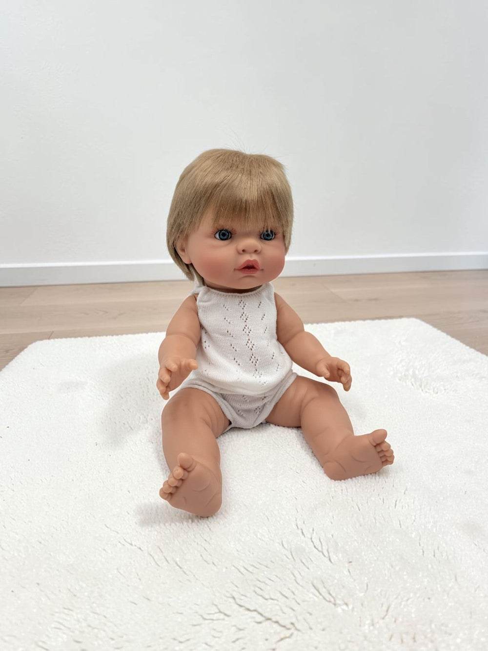 A realistic baby doll with short blonde hair and blue eyes, wearing a white sleeveless knit bodysuit, sitting on a white textured rug in a room with light wood flooring and a white wall.