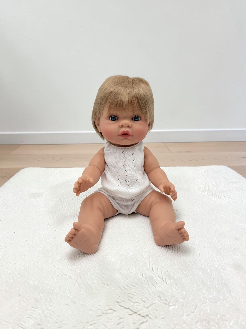 A realistic baby doll with short blonde hair and blue eyes sits on a white textured rug in a room with light wood flooring and a white wall.