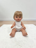 A realistic baby doll with short blonde hair and blue eyes sits on a white textured rug in a room with light wood flooring and a white wall.