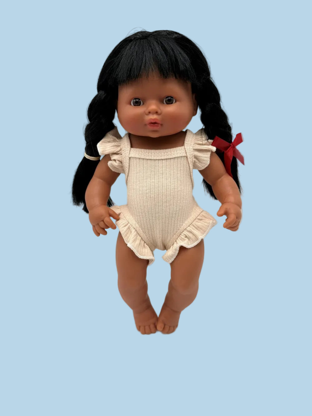 A baby doll with black hair in two braids, wearing a cream ribbed romper with ruffled sleeves and hem, and a red hair tie, standing against a plain white background.
