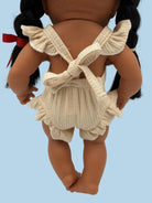 A close-up back view of a doll with dark brown skin and black pigtails, wearing a cream ribbed romper with ruffled straps and a bow tie back, photographed against a plain white background.