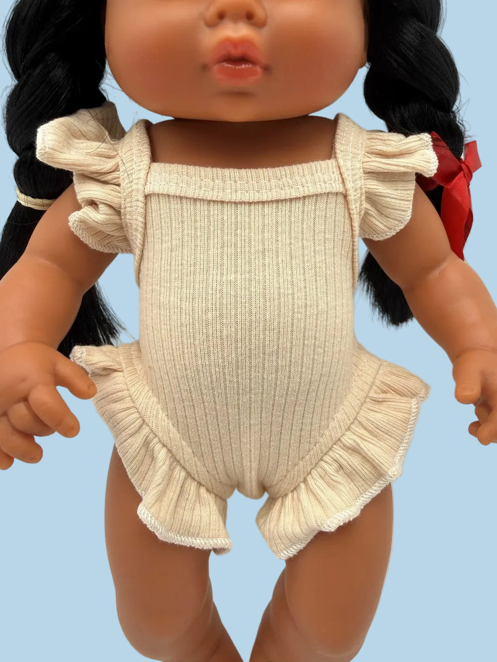 A close-up of a doll wearing a ribbed cream-colored romper with ruffled shoulder straps and a ruffled hem, shown against a plain light background.