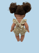 Back view of a doll with brown hair in two buns, wearing a beige and white ruffled romper with a bow on the back, set against a plain light gray background.