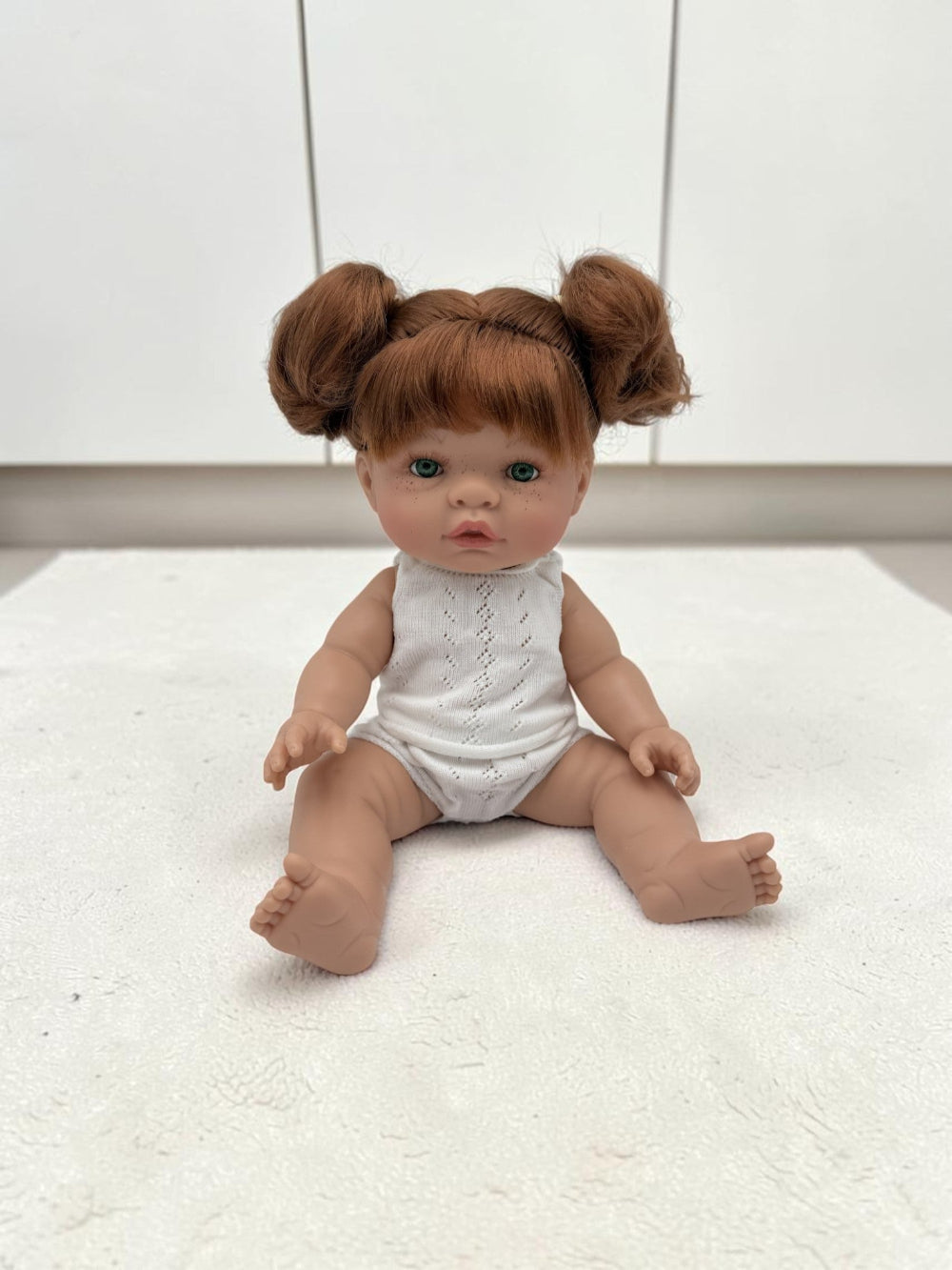 A realistic baby doll with red hair styled in two buns, wearing a white sleeveless top and diaper, sitting on a white textured surface against a white paneled background.