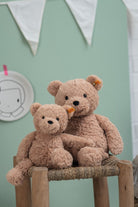 Two teddy bears sitting on a wooden stool against a light green wall.