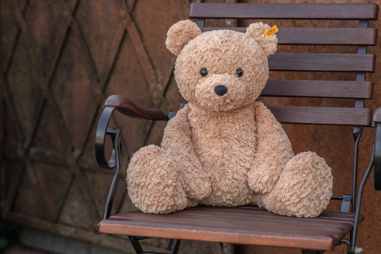 Brown teddy bear sitting on a wooden chair against a textured wall.
