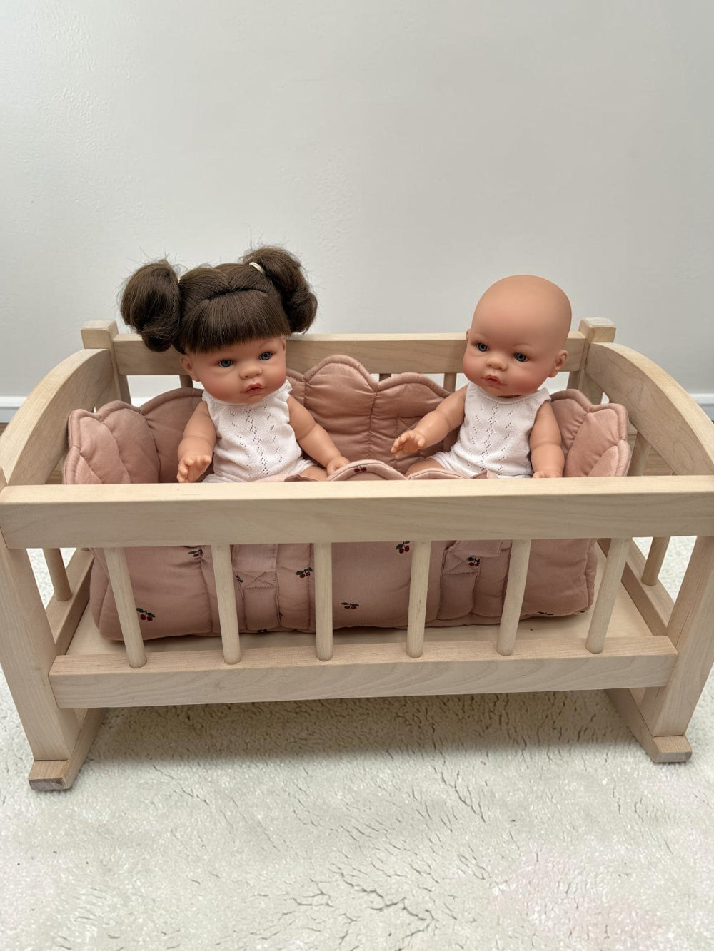 A light natural wood baby cradle with a pink quilted cushion, holding two dolls against a plain light gray wall and white textured floor.