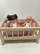 A light natural wood baby cradle with a pink quilted cushion, holding two dolls against a plain light gray wall and white textured floor.