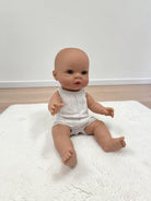 A realistic baby doll with a bald head and blue eyes, wearing a white sleeveless knit romper, sitting on a white textured rug against a plain white wall and light wood floor.