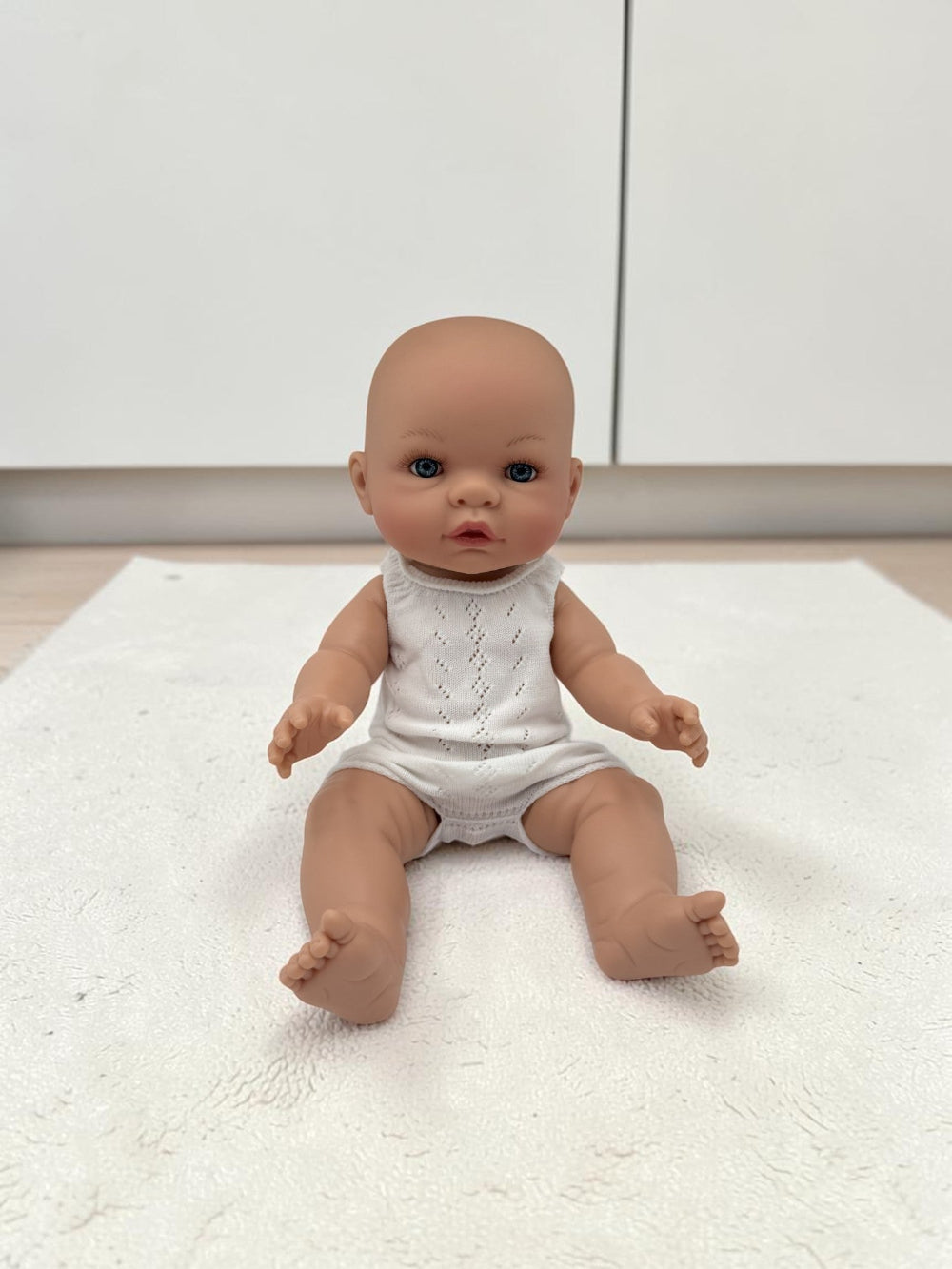 A realistic baby doll with a bald head and blue eyes, wearing a white sleeveless romper, sitting on a white textured rug in front of white cabinets.