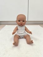 A realistic baby doll with a bald head and blue eyes, wearing a white sleeveless romper, sitting on a white textured rug in front of white cabinets.