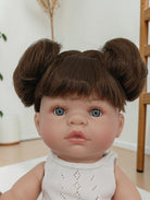 Realistic baby doll with brown hair styled in buns, wearing a white sleeveless top.