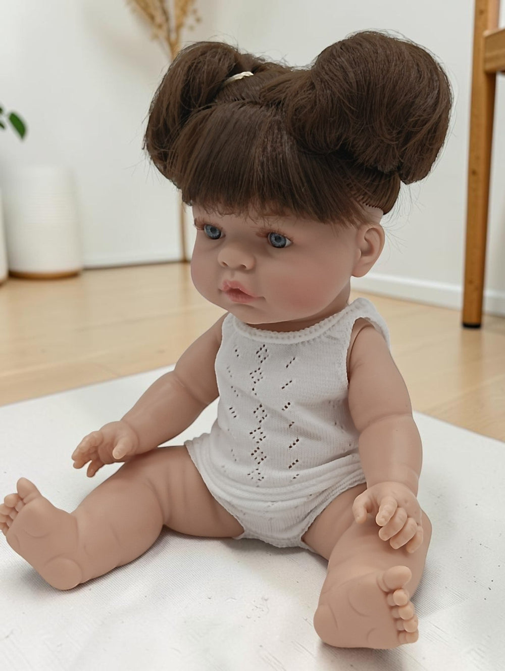 Realistic baby doll with brown hair styled in buns, wearing a white lace romper, on a light-colored surface.
