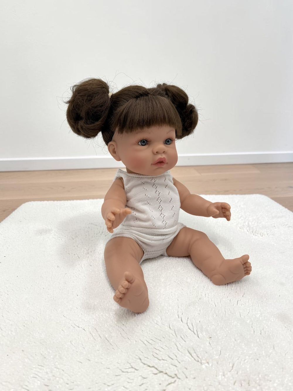 A realistic baby doll with brown hair in two buns sits on a white textured rug in a room with light wood flooring and a white wall.