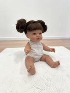 A realistic baby doll with brown hair in two buns sits on a white textured rug in a room with light wood flooring and a white wall.