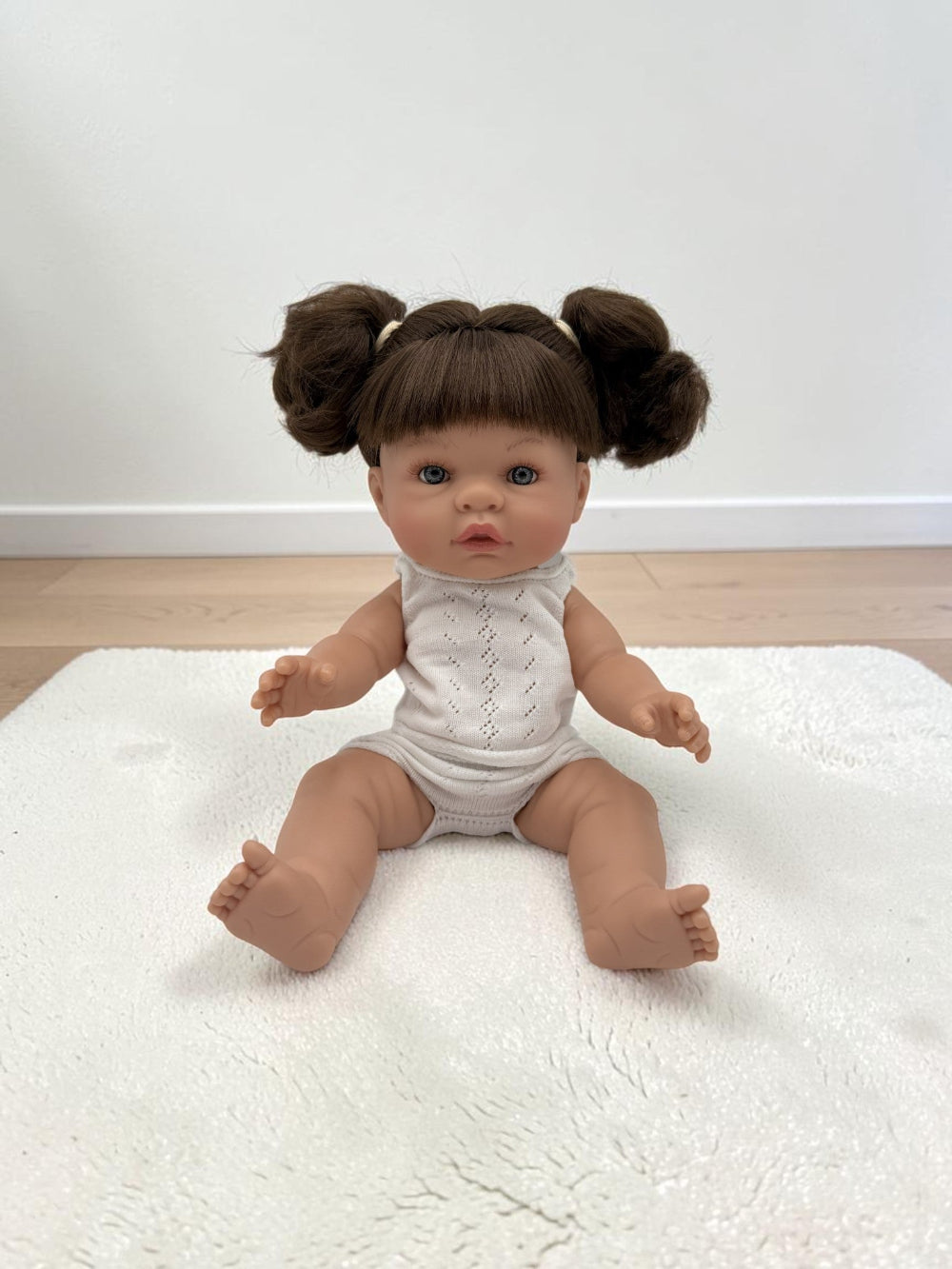 A realistic baby doll with brown hair in two buns sits on a white textured rug in a room with a white wall and light wood floor. The doll is wearing a white sleeveless knit bodysuit.