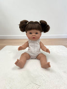 A realistic baby doll with brown hair in two buns sits on a white textured rug in a room with a white wall and light wood floor. The doll is wearing a white sleeveless knit bodysuit.