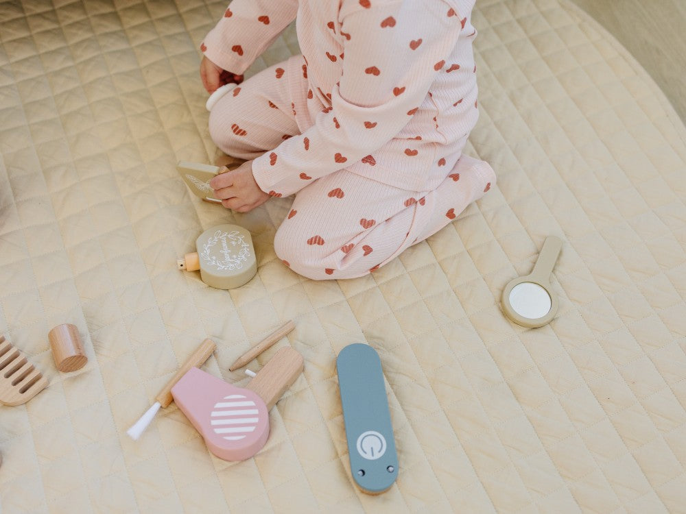 Child in pink pajama with heart patterns sitting on a bed surrounded by wooden toys.