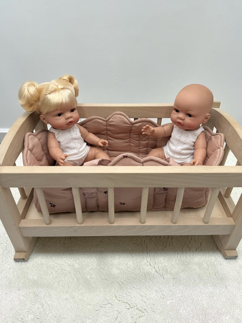 A miniature light wood doll crib with a pink quilted cushion, holding two baby dolls dressed in white outfits, set against a plain light gray wall and textured floor.
