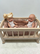 A miniature light wood doll crib with a pink quilted cushion, holding two baby dolls dressed in white outfits, set against a plain light gray wall and textured floor.