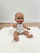A baby doll with realistic skin and dark eyes sits on a white textured rug in a room with light wood flooring and a white wall.