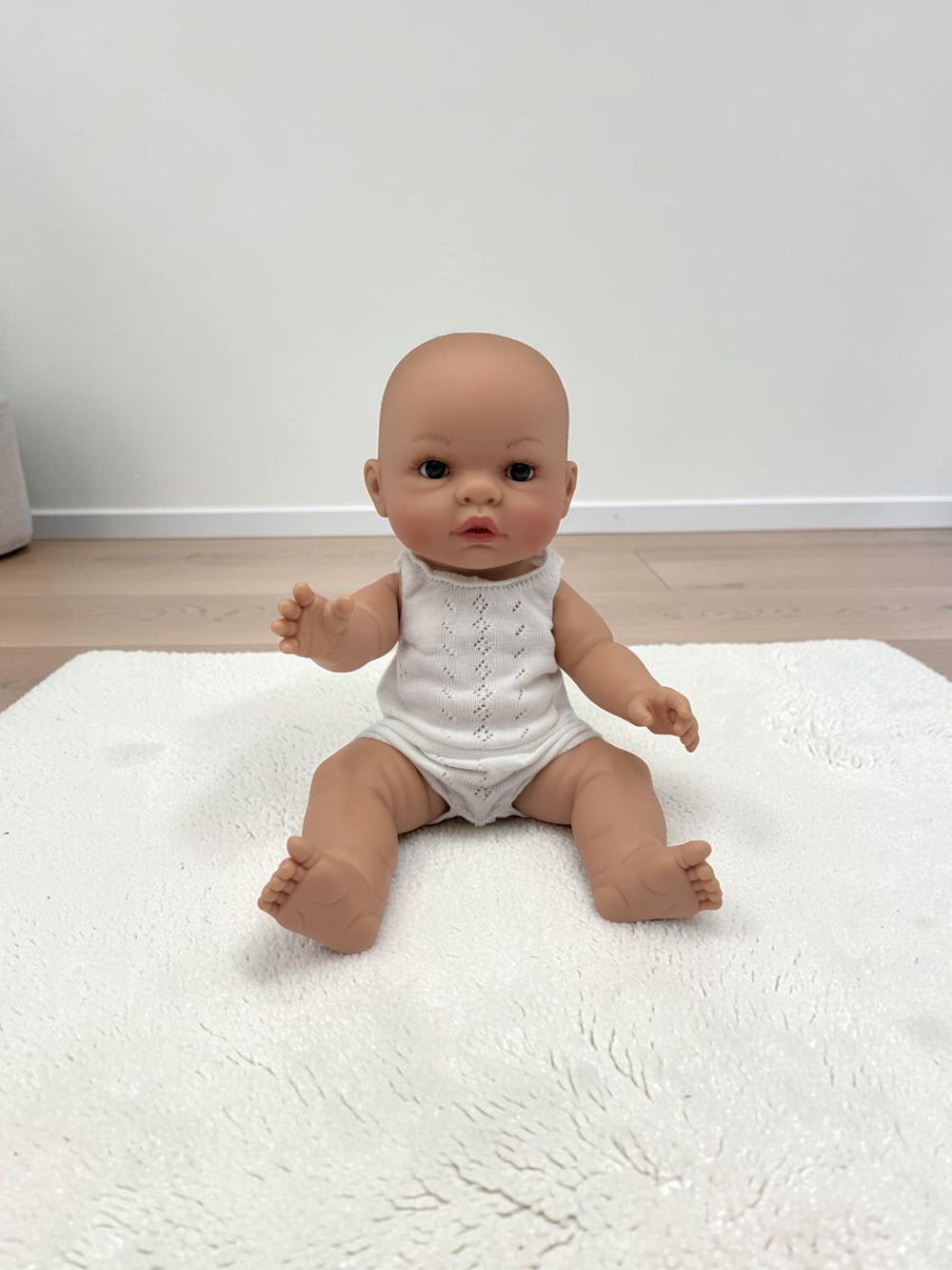 A realistic baby doll with a bald head, realistic skin tone, and dark eyes, wearing a white sleeveless bodysuit, sitting on a white textured rug in a room with light wood flooring and a white wall.