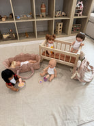 A doll play set arranged on a light beige fabric floor in front of a wooden toy storage shelf. The set includes a light wood baby crib with two dolls, a pink stroller with a doll lying inside, a pink bunny backpack, a rocking horse, and small toy accessories.